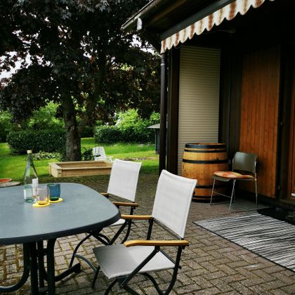 Ein gemütlicher Außenbereich mit einem Tisch und Stühlen. Im Hintergrund sieht man einen gepflegten Garten mit grüner Vegetation.