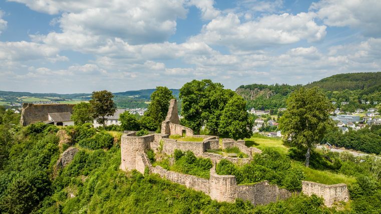 Ruinen der Löwenburg in Gerolstein, umgeben von grüner Landschaft und Bäumen, unter blauem Himmel mit Wolken.