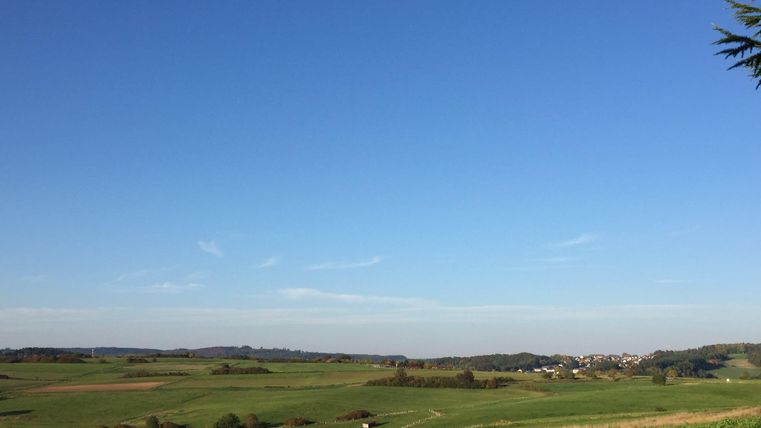 Eine weite Landschaft mit grünen Feldern und klarem blauem Himmel. In der Ferne sind sanfte Hügel und ein kleines Dorf zu sehen.