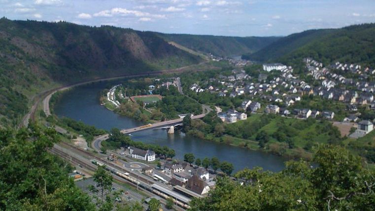 Eine malerische Landschaft mit einem Fluss, der durch grüne Hügel fließt. Im Hintergrund sind kleine Häuser und eine Brücke zu sehen.