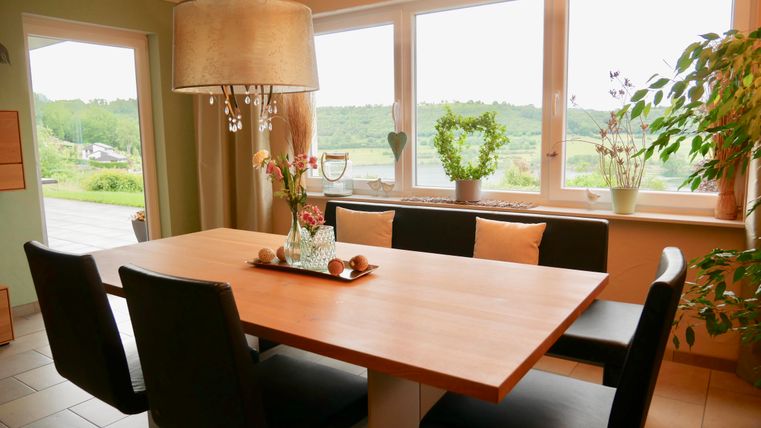 Ein helles Esszimmer mit einem großen Holztisch und schwarzen Stühlen. Durch die Fenster sieht man eine grüne Landschaft mit Pflanzen und Dekorationen im Raum.