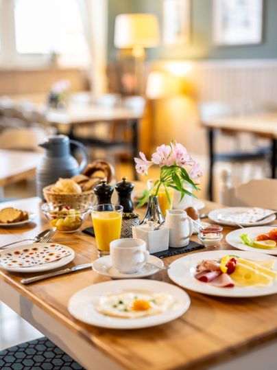 Ein reichhaltiges Frühstück auf einem Holztisch mit frischen Brötchen, Obst und Getränken. Eine hübsche Blume in einer Vase sorgt für eine einladende Atmosphäre.