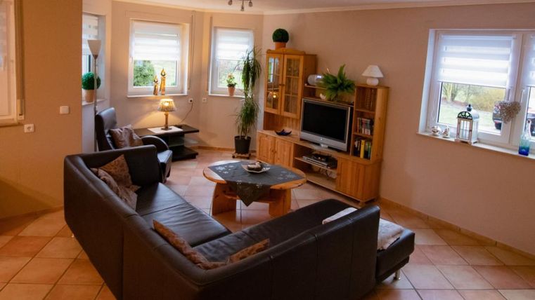 Ein gemütliches Wohnzimmer mit einer großen, dunklen Couch und einem Tisch in der Mitte. Es gibt Fenster mit Licht und eine Holzmöbelkommode, die einen Fernseher beherbergt.