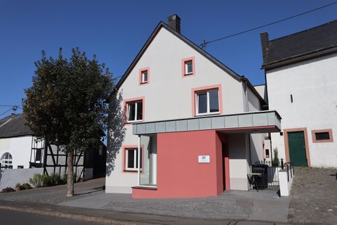 Ein modernes Haus mit einem pinken Anbau und großen Fenstern. Die Umgebung ist ruhig mit Bäumen und anderen Gebäuden.