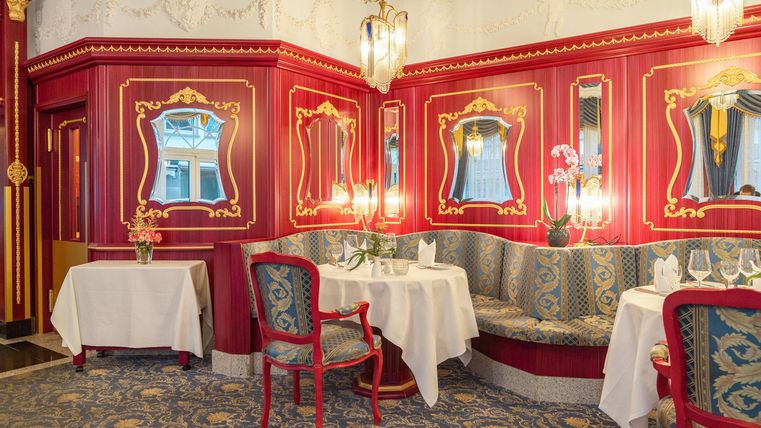 Ein elegantes Restaurant mit rotem Wandanstrich und stilvollen Spiegeln. Gemütliche Tische und ein geschmackvolles Ambiente laden zum Verweilen ein.