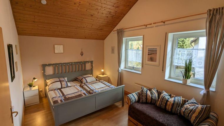 Ein gemütliches Schlafzimmer mit einem Doppelbett und einem Sofa. Die Wände sind hell, und große Fenster lassen viel Licht herein.