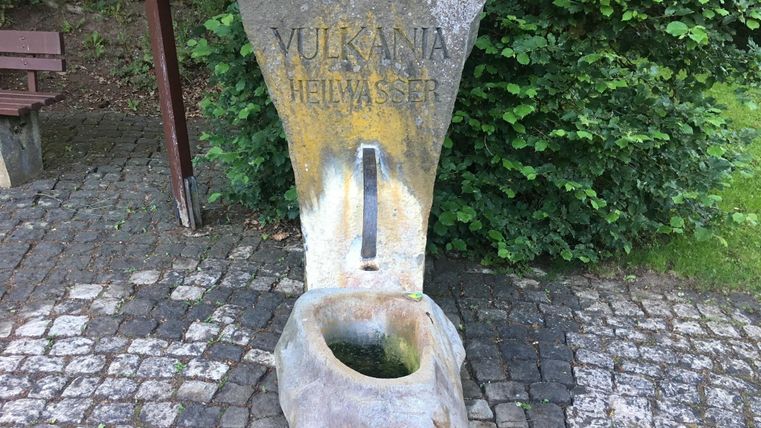 Ein Steinspringbrunnen mit der Aufschrift „Vulkana Heilwasser“. Der Brunnen ist von grünen Pflanzen umgeben und steht auf einem gepflasterten Weg.
