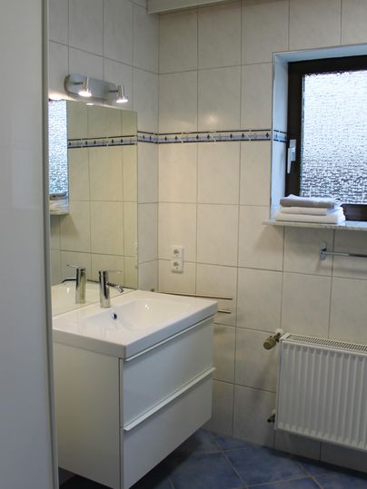 Ein modernes Badezimmer mit einem Doppelwaschtisch und einem großen Spiegel. Die Wände sind hell gefliest, und ein Fenster sorgt für natürliches Licht.