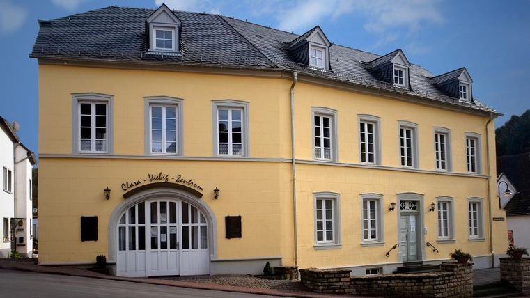 Ein gelbes Gebäude mit grauem Dach und weißen Fenstern. Es gibt eine große Eingangstür und eine ruhige Atmosphäre.