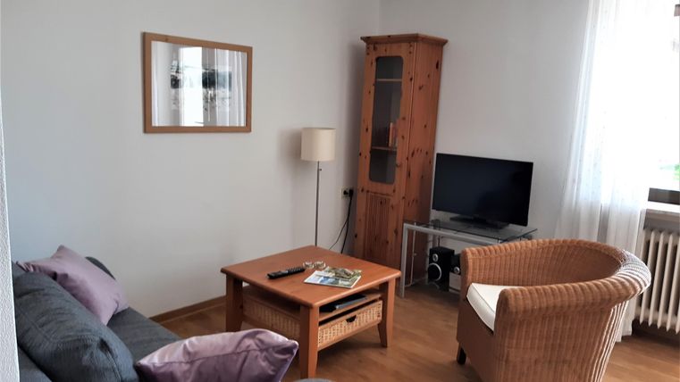 Ein gemütliches Wohnzimmer mit einem Sofa, einem Sessel und einem Holztisch. Es gibt einen Fernseher und viel Licht durch das Fenster.