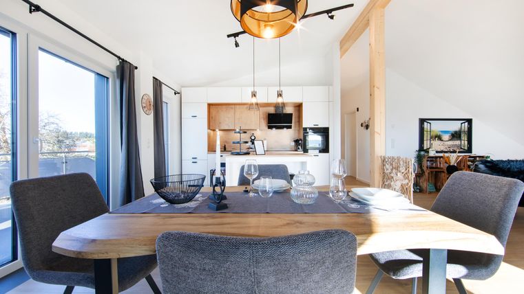 A modern dining room with a large wooden table and stylish chairs. In the background, the open kitchen is in bright colors,