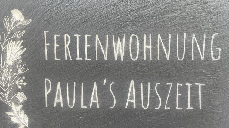 Ein Schild mit der Aufschrift "Ferienwohnung Paula's Auszeit". Es zeigt eine einladende Atmosphäre für Urlaubsgäste.