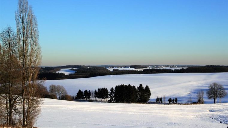 Eine schneebedeckte Landschaft mit weitem Blick auf Felder und Bäume. Der Himmel ist klar und blau.