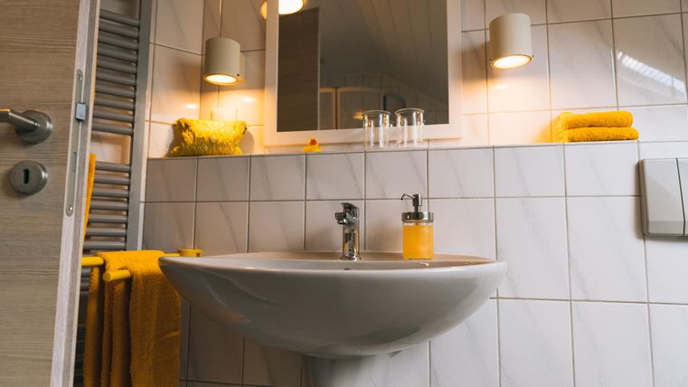 Ein modernes Badezimmer mit einem weißen Waschbecken und einem großen Spiegel. Gelbe Handtücher und dekorative Elemente sorgen für eine gemütliche Atmosphäre.