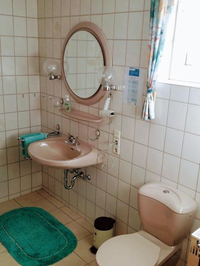 Ein sauberes Badezimmer mit einer Toilette und einem Waschbecken. Auf dem Boden liegen grüne Badeteppiche.