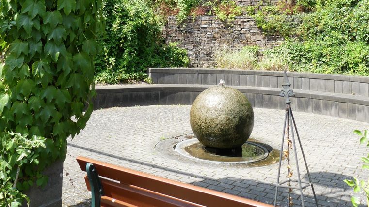 Ein ruhiger Garten mit einer runden Wasserfontäne in der Mitte. Eine Holzbank lädt zum Verweilen ein, um die grüne Umgebung zu genießen.