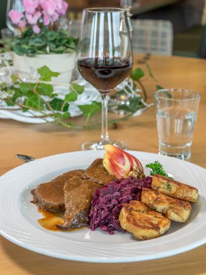 Ein Teller mit zartem Fleisch, Rotkohl und Kartoffelklößen. Im Hintergrund steht ein Glas Rotwein und ein Wasserglas.
