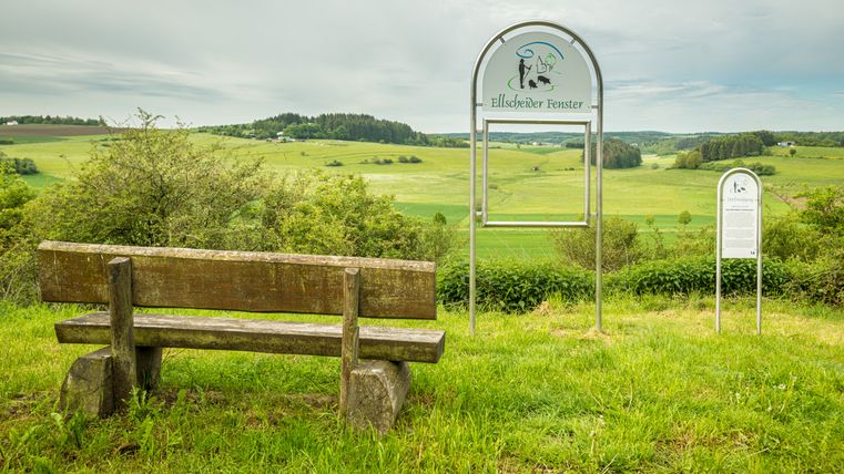 Houten bank en bord met de inscriptie 'Ellscheider Fenster' voor een groen landschap.