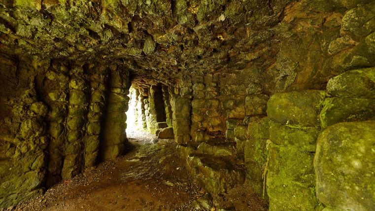 Ein schmaler, von Felsen umgebener Tunnel mit moosbedeckten Wänden. Am Ende des Tunnels ist helles Licht sichtbar.