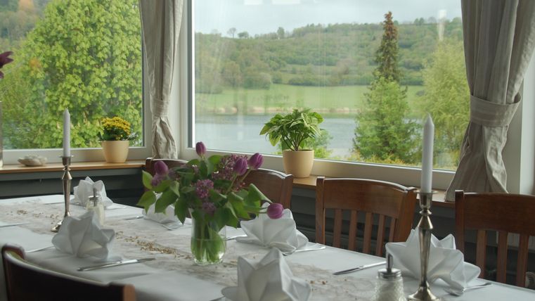 Ein schön gedeckter Tisch mit Blumenarrangement und Kerzen. Durch das Fenster ist eine grüne Landschaft mit einem Gewässer sichtbar.