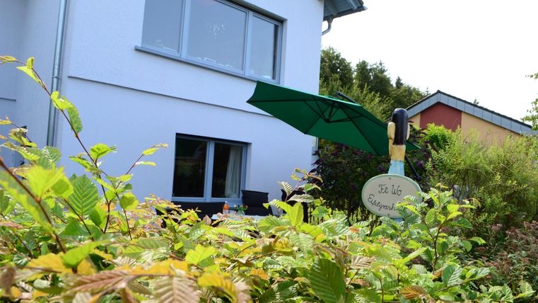 Ein modernes Haus mit einem grünen Sonnenschirm und einem Gartenschild. Vorne wachsen verschiedene Pflanzen und Sträucher.