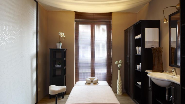 Ein ruhiger Wellnessraum mit einem Massagetisch und sanften Farben. Es gibt Fenster mit Jalousien und eine elegante Dekoration.