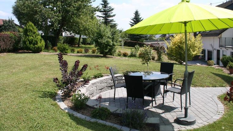 Ein einladender Garten mit einer runden Steinterrasse, einem Tisch und Stühlen. Ein großer, leuchtend grüner Sonnenschirm bietet Schatten.