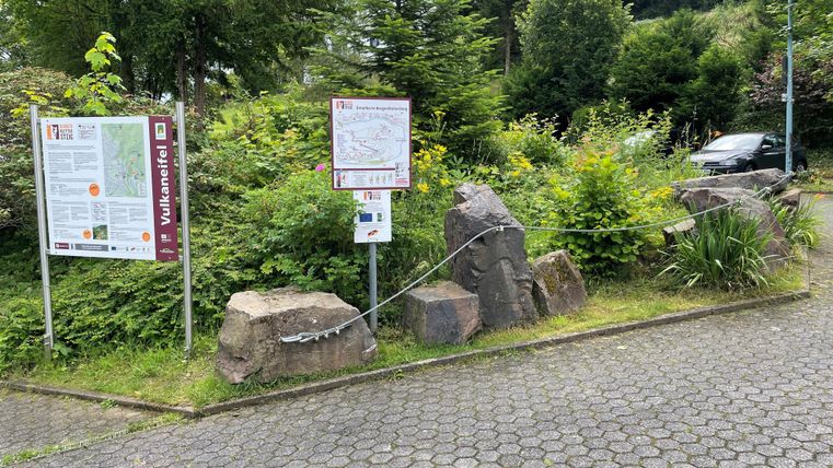 Ein Eingangsbereich mit Infotafeln und Steinen, umgeben von grünen Sträuchern und Bäumen. Im Hintergrund ist ein Parkplatz zu sehen.