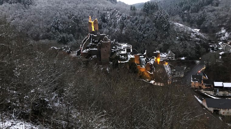 Een besneeuwd landschap met een historisch kasteel op een heuvel. Op de achtergrond zijn beboste bergen zichtbaar.
