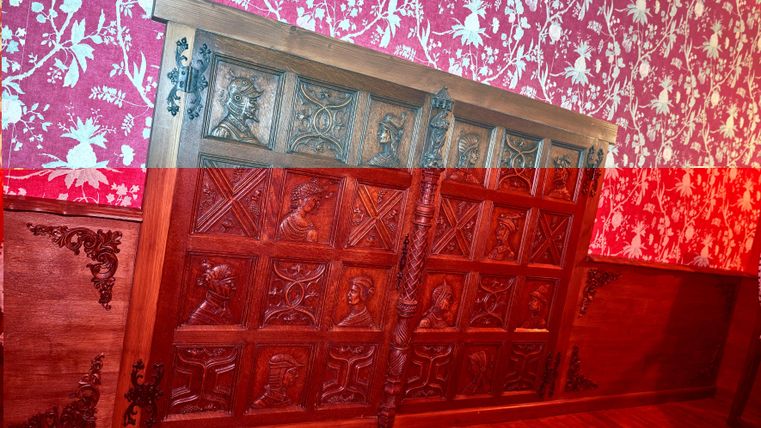 Ein kunstvoll geschnitztes Holzpaneel mit verschiedenen Reliefs. Es ist an einer Wand mit rot-weißer Blumentapete angebracht.
