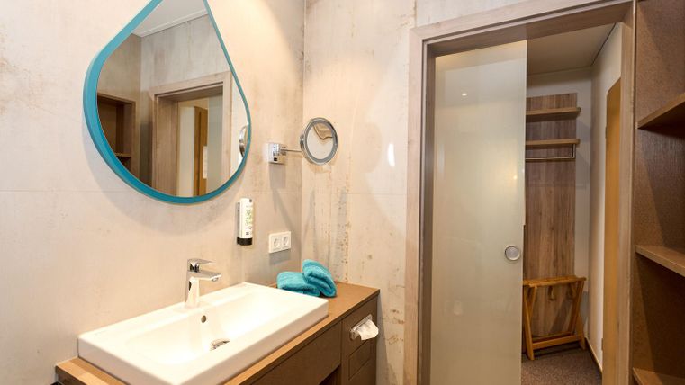 Ein modernes Badezimmer mit einem weißen Waschbecken und einem großen, runden Spiegel. Der Raum ist hell und minimalistisch gestaltet, mit einem Holzregal und einer Duschkabine.