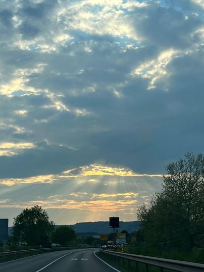 Eine Straße, die durch eine ländliche Umgebung führt, unter einem bewölkten Himmel. Sonnenstrahlen brechen sanft durch die Wolken kurz vor dem Sonnenuntergang.