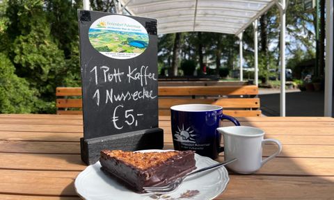 Ein Stück Nussschokoladenkuchen auf einem Teller, neben einer Tasse Kaffee und einer kleinen Kanne. Im Hintergrund steht ein Schild mit dem Angebot: "1 Pott Kaffee 1 Nussecke €5".