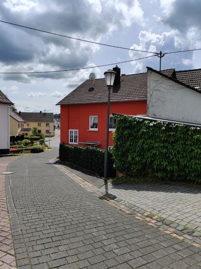Eine charmante Straße mit einem roten Haus und gepflastertem Boden. Im Hintergrund sind Wolken und einige Häuser zu sehen.