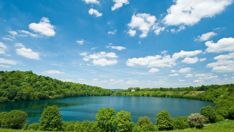 Ein schöner See umgeben von grünen Hügeln und Bäumen. Der Himmel ist klar und blau mit einigen Wolken.