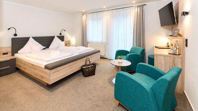 Ein modernes Hotelzimmer mit einem Doppelbett, einer gemütlichen Sitzecke und einem Fernseher. Große Fenster sorgen für viel Tageslicht und eine angenehme Atmosphäre.