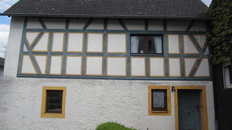 Ein traditionelles Fachwerkhaus mit einer weißen Fassade und blauen Balken. Der Eingang ist grün und von einem kleinen Strauch umgeben.