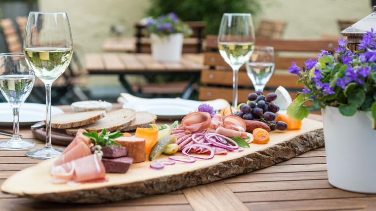 Eine ansprechende Platte mit verschiedenen Wurst- und Käsesorten, Obst und Gemüse. Im Hintergrund stehen Gläser mit Weißwein und eine blühende Pflanze auf einem Tisch im Freien.