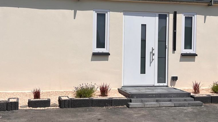 Ein modernes Haus mit einer weißen Eingangstür und großen Fenstern. Vor dem Haus befinden sich gepflegte Pflanzen und ein schöner Weg.