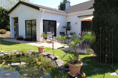 Ein schöner Garten mit einem Teich und bunten Pflanzen. Das Haus hat große Fenster und eine gemütliche Terrasse.