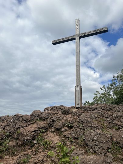 Ein hölzernes Kreuz steht auf einem Felsen unter einem bewölkten Himmel. Grüne Pflanzen umgeben die felsige Umgebung.