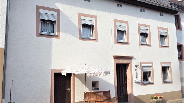 Ein einfaches, zweigeschossiges Haus mit weißen Wänden und einem roten Rahmen. Vor dem Haus steht eine Bank und ein Sonnenschirm.