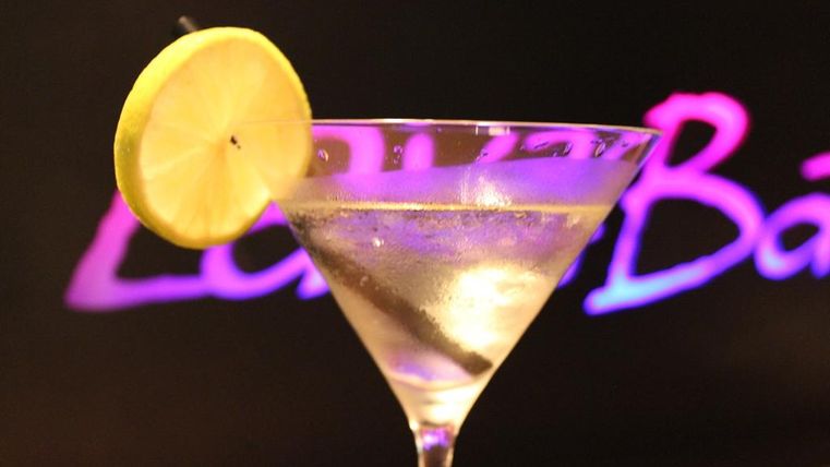 Ein Cocktail in einem Martini-Glas mit einer Zitronenscheibe als Garnitur. Im Hintergrund leuchtet ein bunter Schriftzug.