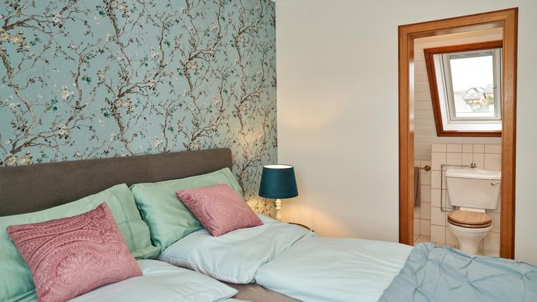 Ein gemütliches Schlafzimmer mit einem modernen Bett und bunten Kissen. Im Hintergrund ist ein hellblauer Wandteppich und ein Badezimmer sichtbar.