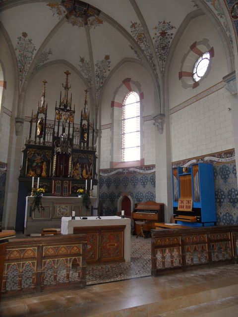 An interior of a church with an artistically designed altar and colorful windows. On the left side stands an organ, and the walls are adorned with historic paintings.