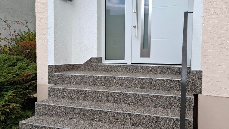 Ein moderner Eingang mit Stufen und einer gläsernen Haustür. Die Umgebung ist gepflegt und einladend.