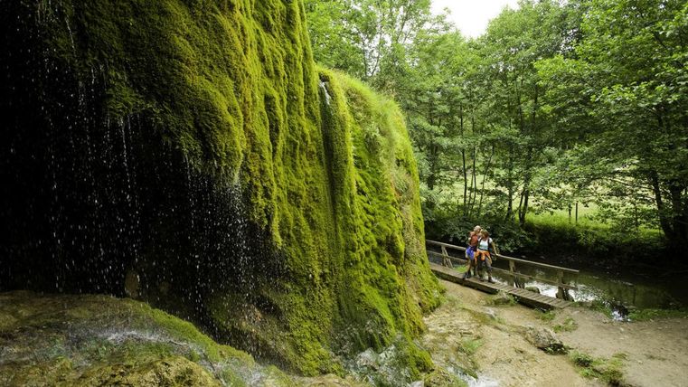 Ein grüner, moosbedeckter Felsen mit plätscherndem Wasser. Im Hintergrund sind Bäume und ein Wanderer auf einem Steg zu sehen.