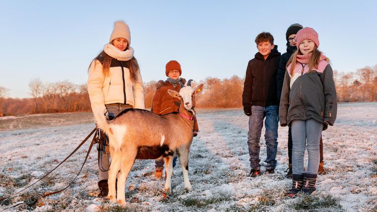 Eine Gruppe von vier Kindern steht fröhlich auf einem verschneiten Feld. Sie halten eine Ziege an der Leine und genießen den winterlichen Tag.