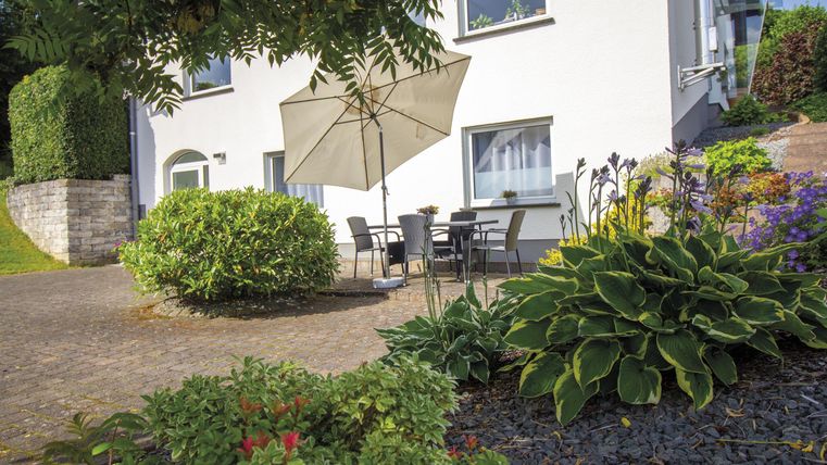 Ein gemütlicher Außenbereich mit einem Tisch, Stühlen und einem Sonnenschirm. Umgeben von bunten Pflanzen und Sträuchern.