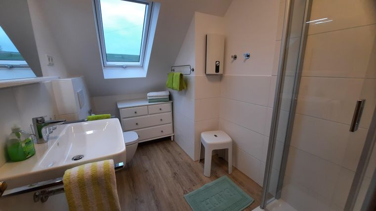 Ein modernes Badezimmer mit einer Dusche und einem Waschbecken. Es gibt helle Wände, Holzfußboden und frische Handtücher in grünen Farben.